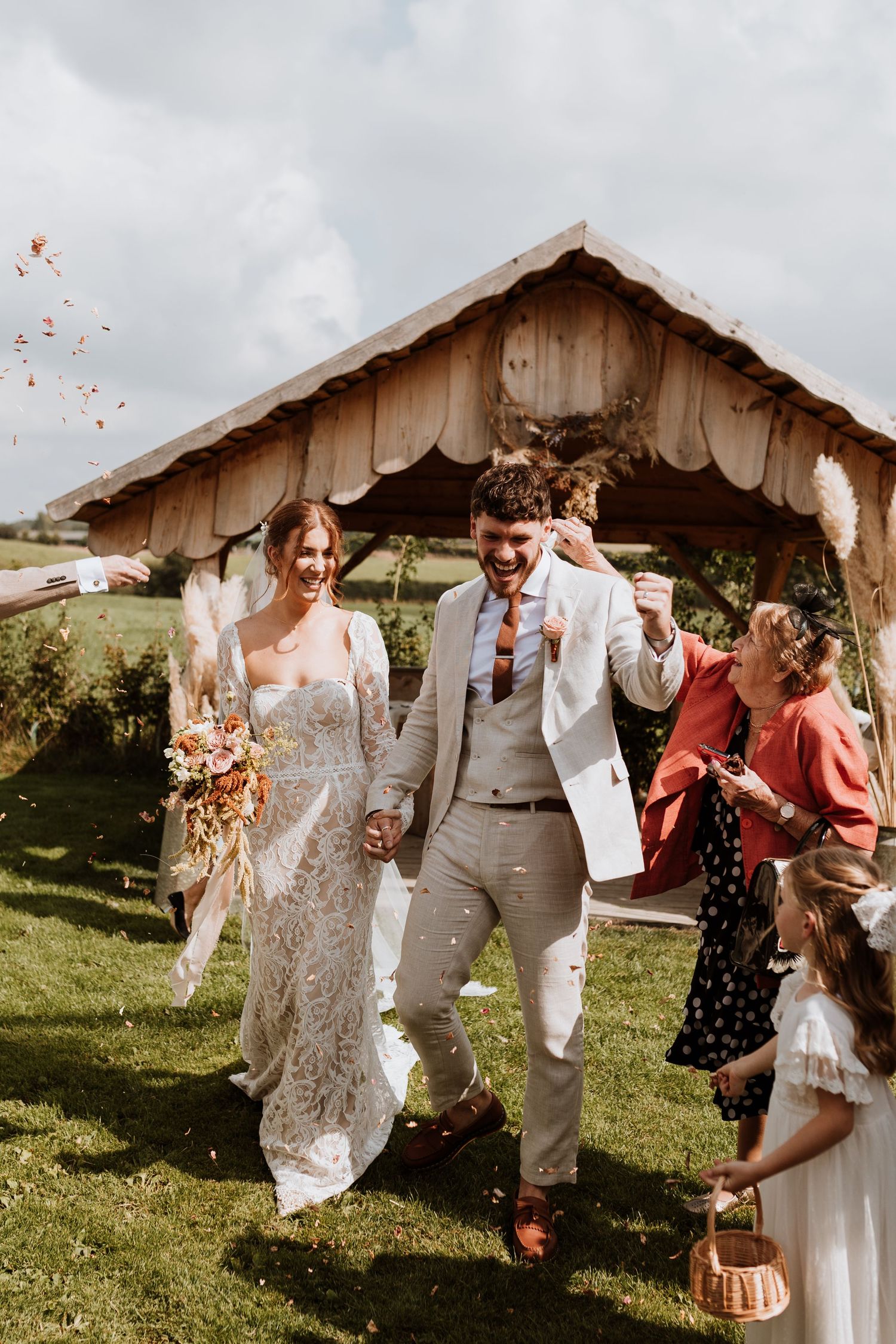 A joyful rustic outdoor wedding celebration with confetti being tossed in front of a wooden barn on a grassy hillside.
