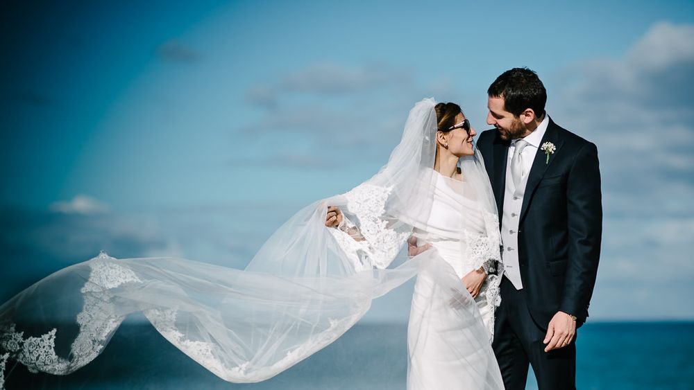 Romantic seaside wedding portraits with flowing veil against blue sky.