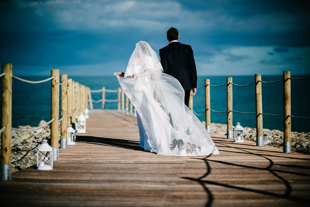 Couple walking on wooden boardwalk by ocean with bride's veil flowing in wind.