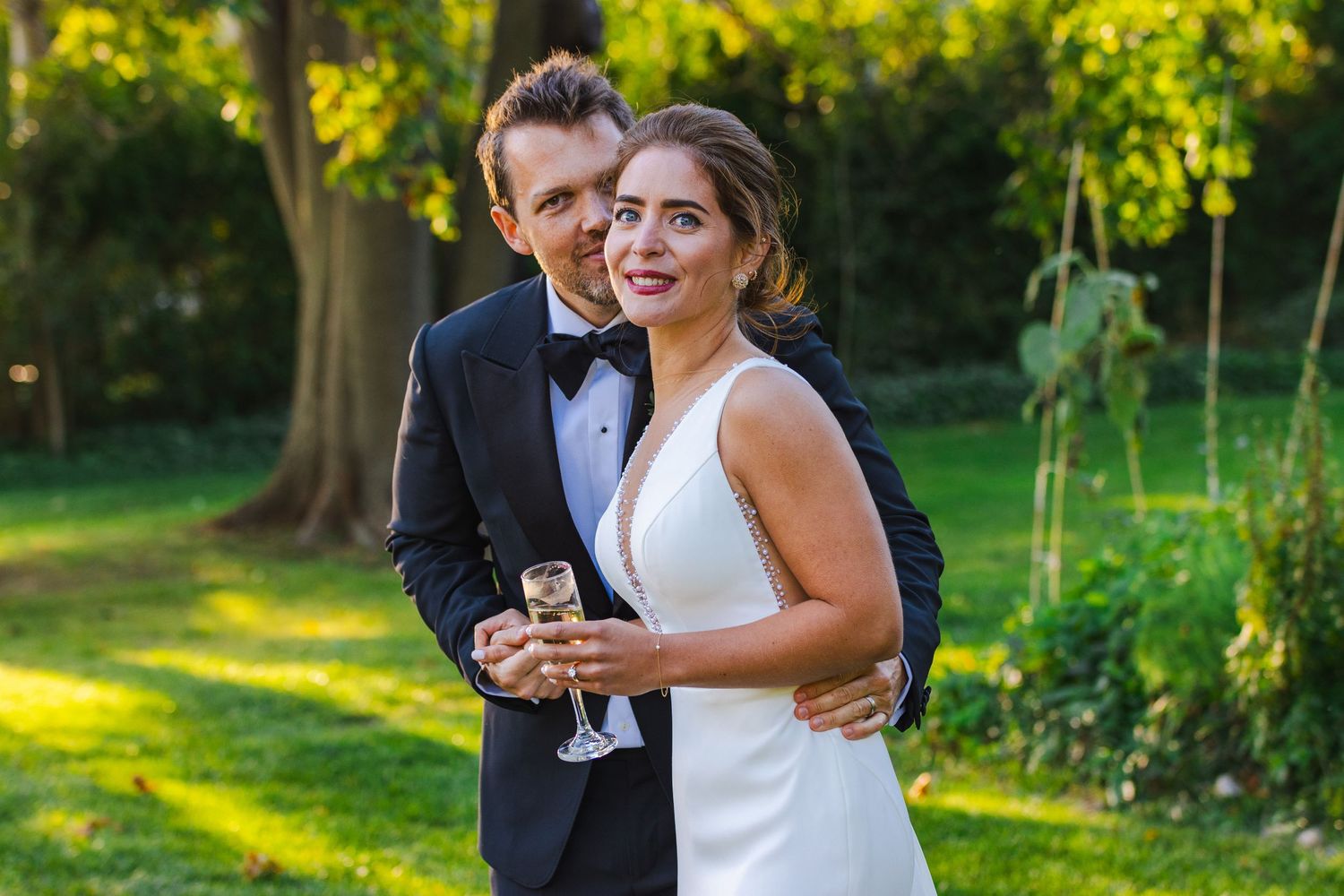 A couple in formal wedding attire sharing a happy moment with wine glasses in a sunlit garden setting.