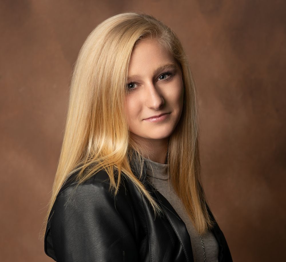 Professional studio portrait of a person with long blonde hair wearing a dark sweater against a brown backdrop.