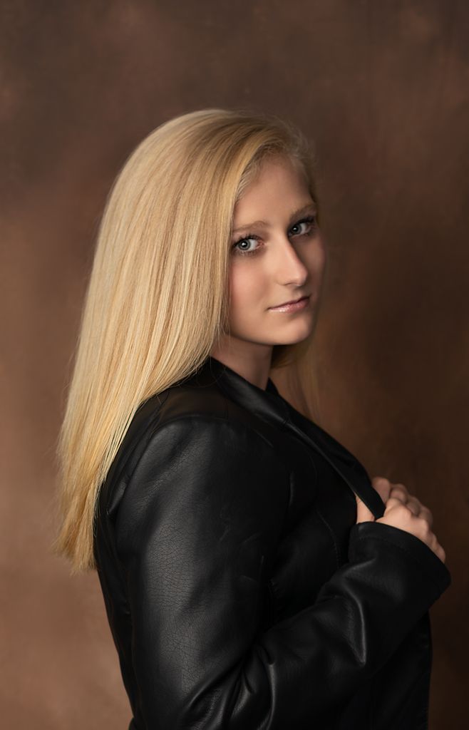 A dramatic studio portrait featuring blonde hair and black clothing against a dark brown background.
