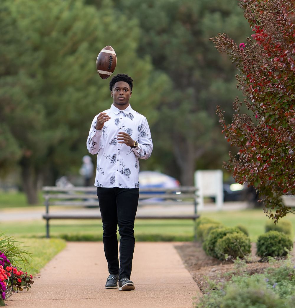 Athletic shot balancing football while walking on pathway with red autumn bushes.