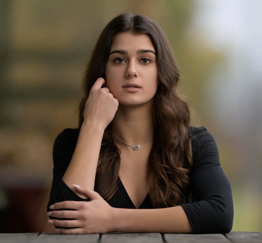 Portrait of person in black sweater with delicate necklace against blurred background.