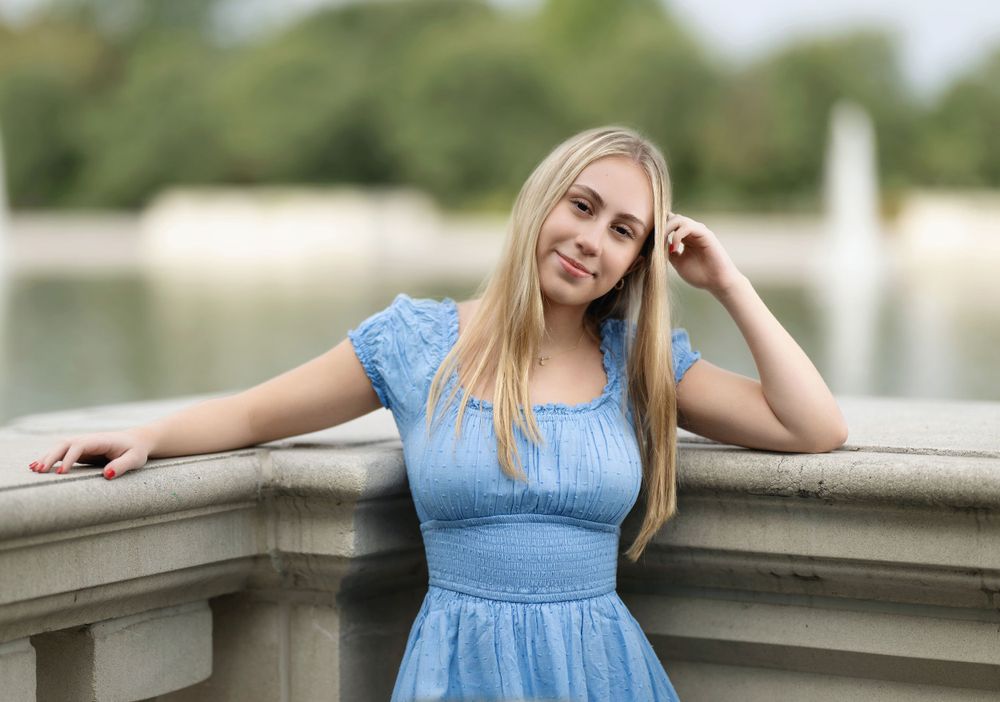 Person in blue dress leaning on stone railing near water during outdoor photoshoot.