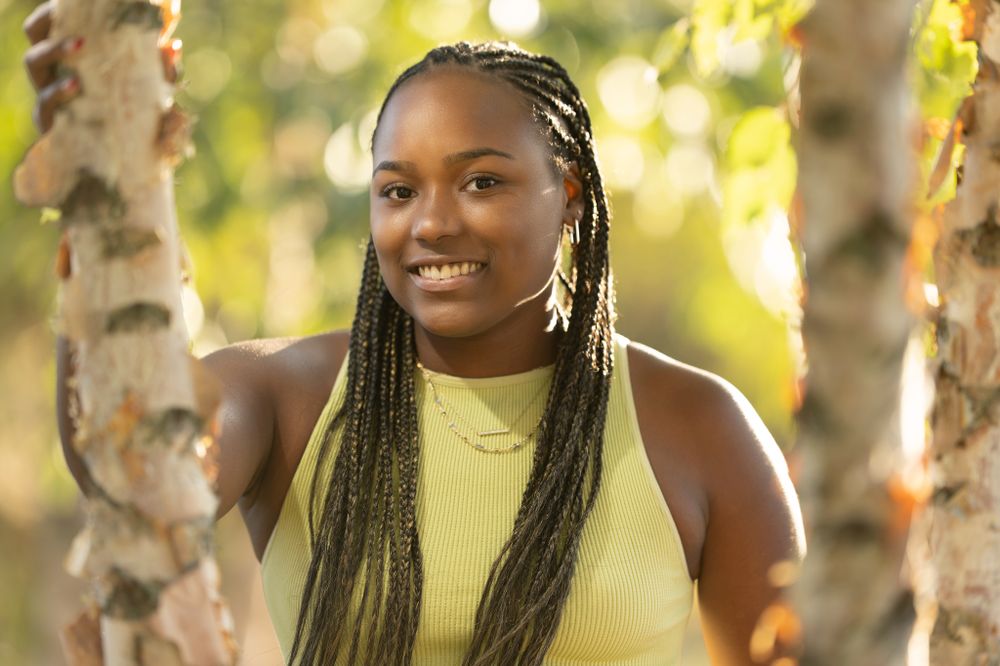 Outdoor portrait of woman with long braids wearing yellow top against sunlit trees.