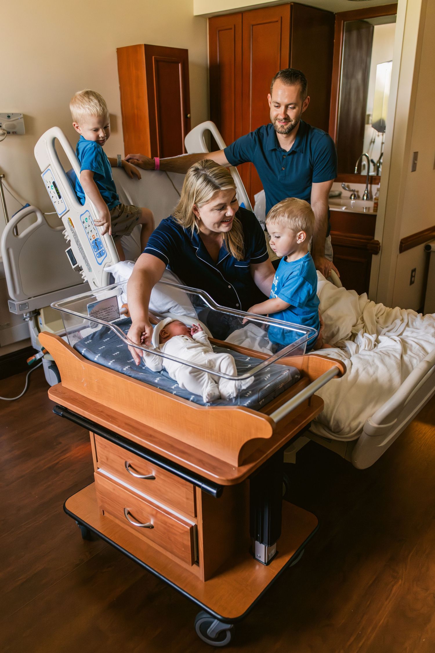 A stainless steel hospital bassinet with a newborn baby inside sits near a bright window.