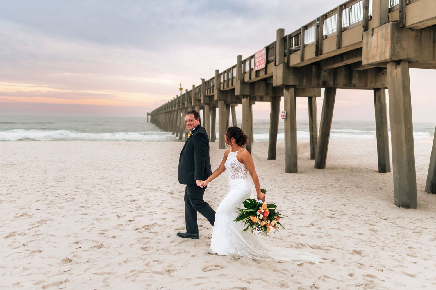Newlyweds walking along beach pier at sunset with bride holding vibrant bouquet.