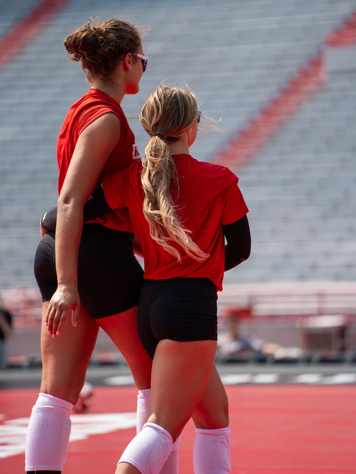 Two athletes in red uniforms warm up on a volleyball court with stadium seating in background.