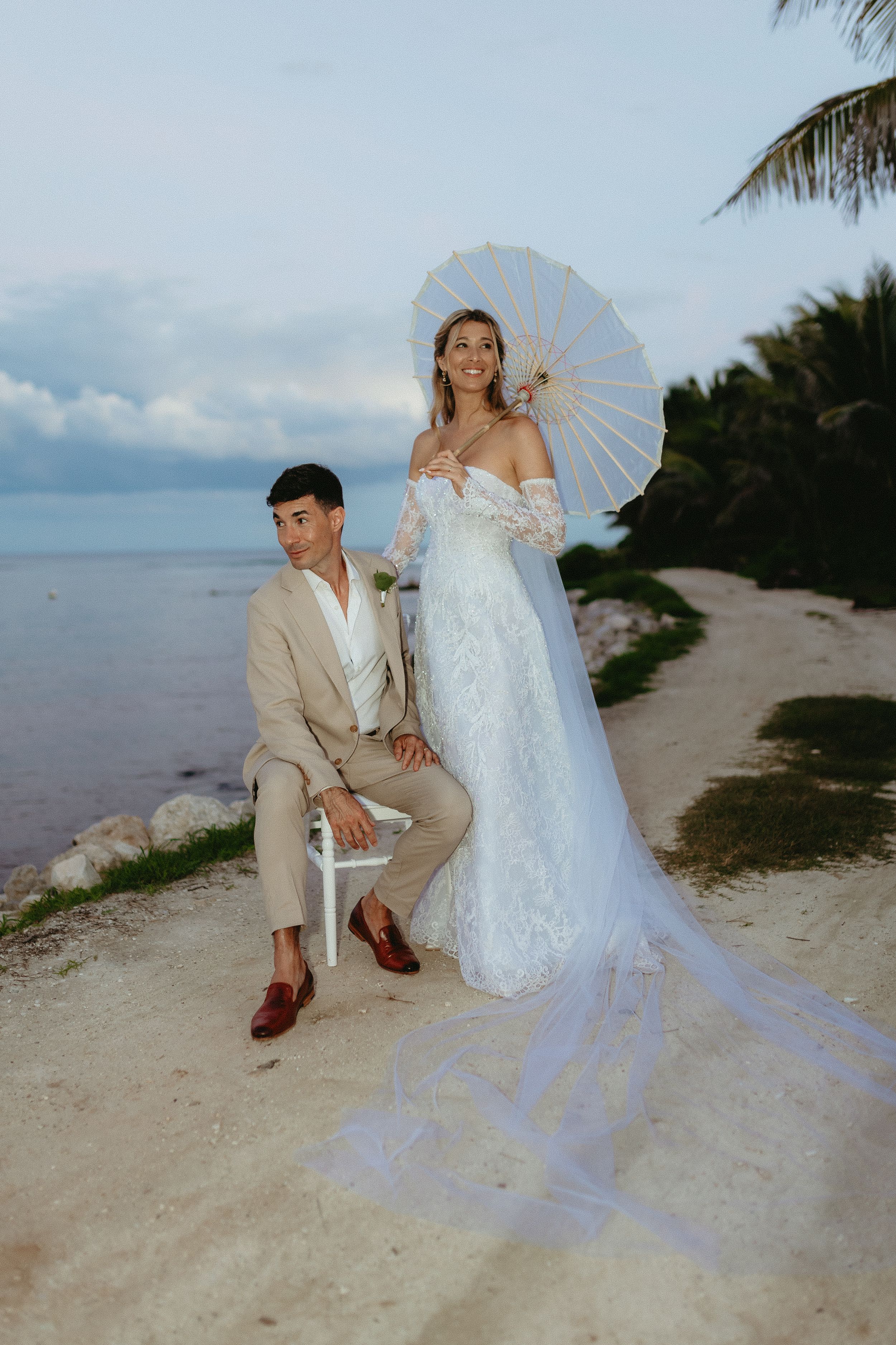 A romantic beach wedding photo with a couple posing on the sand, featuring a white parasol and flowing lace gown.
TRS Coral wedding