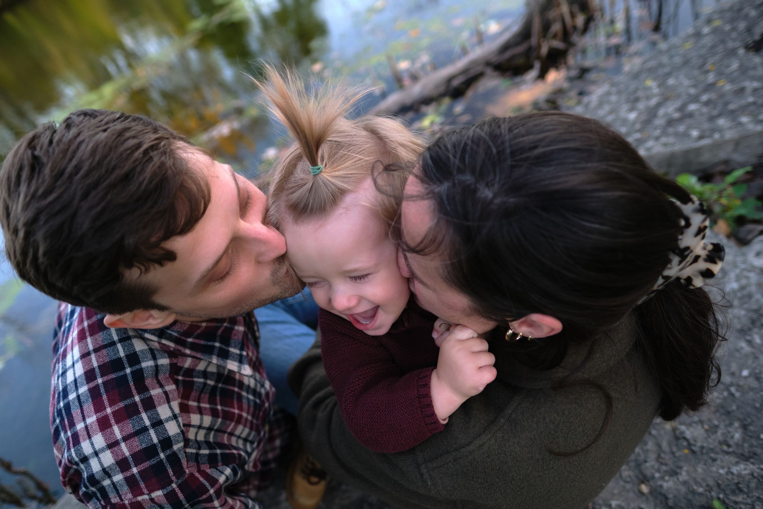 A family portrait of two parents giving their child a kiss on the cheek while sitting near a lake