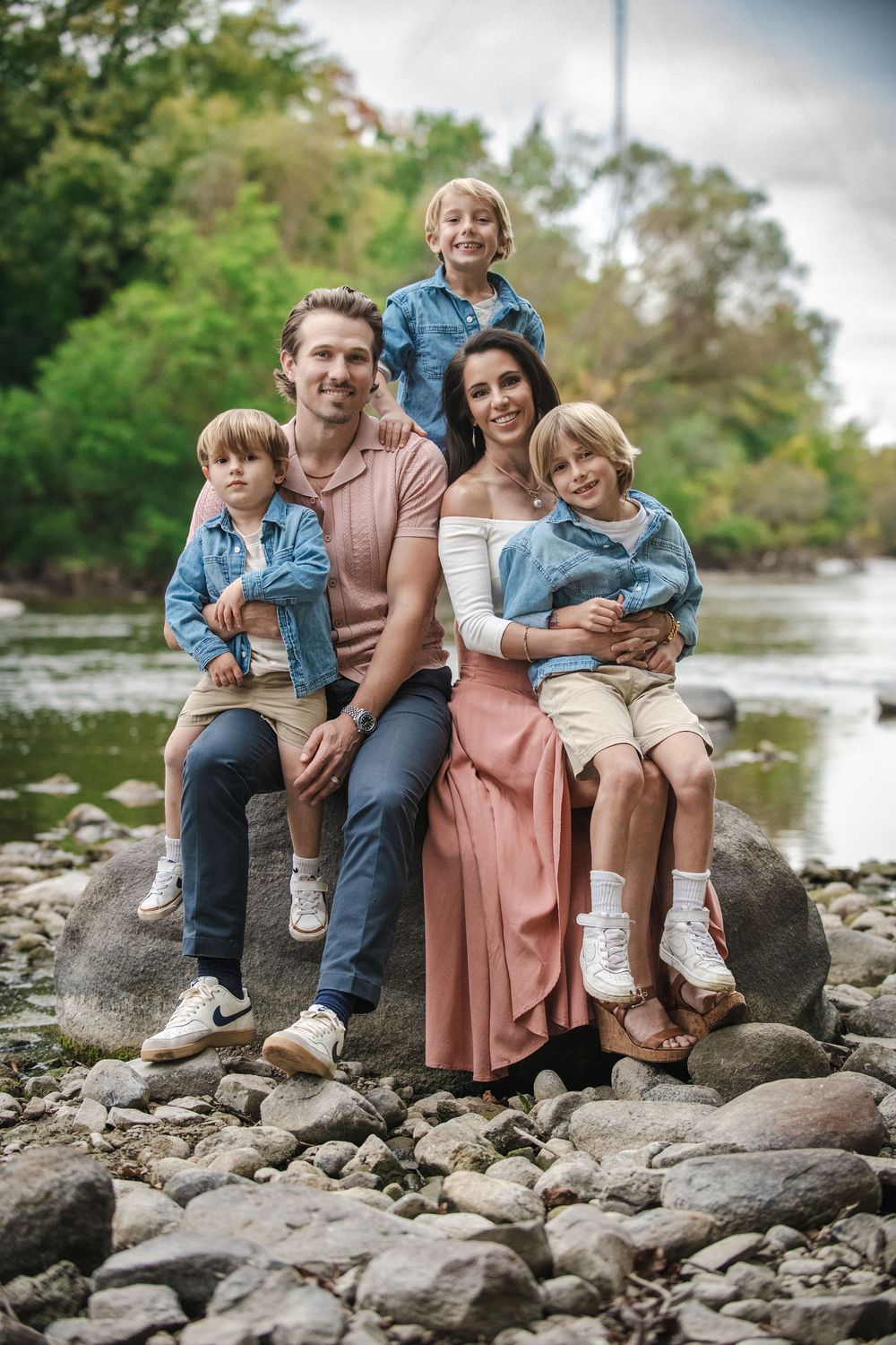 Happy family of five posing on rocks by a river, parents with three young children in denim jackets outdoors.