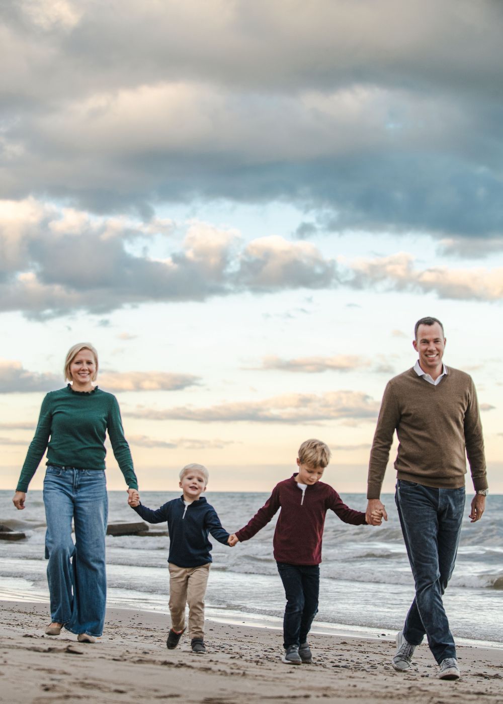 Family of four walking hand-in-hand on a beach at sunset, with cloudy skies and ocean waves in the background.