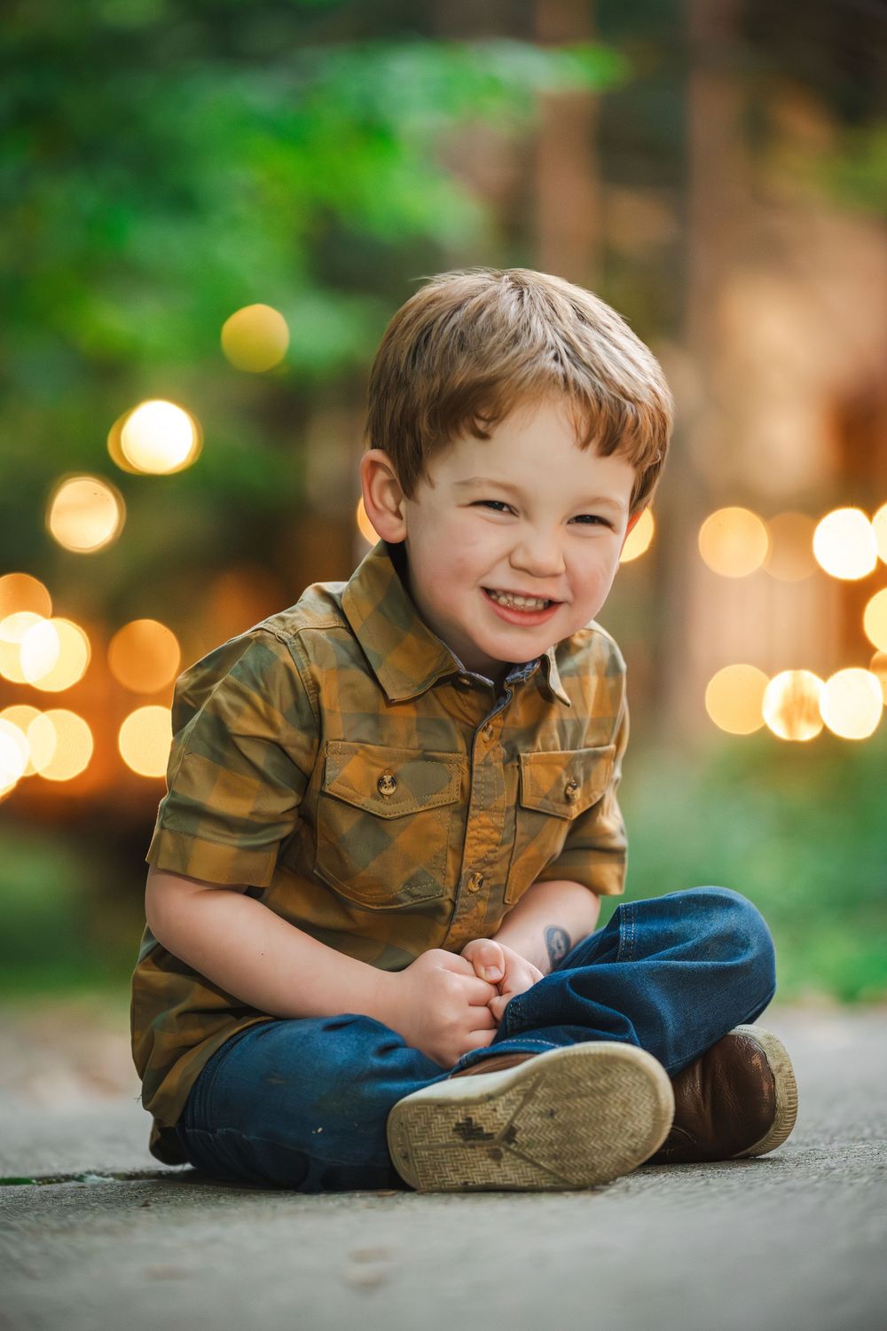 Smiling young boy in plaid shirt and jeans sitting cross-legged outdoors with bokeh lights in background.