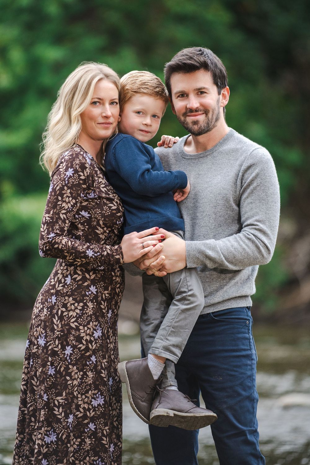 A happy family of three poses outdoors, mom in floral dress, dad in gray sweater holding their young blond son.