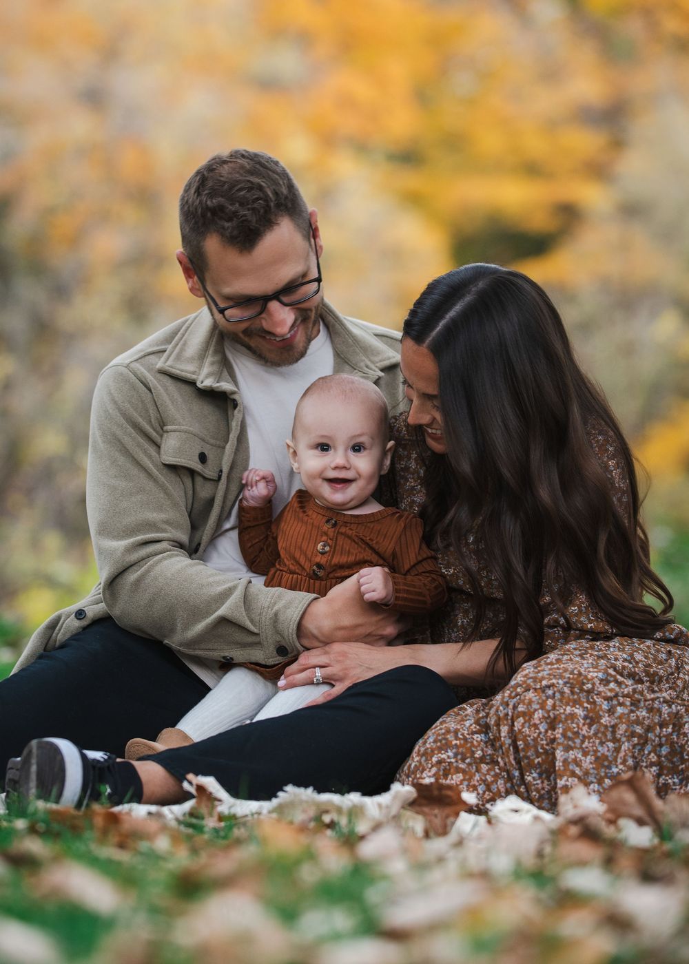 Family of three sitting outdoors in autumn, parents smiling at happy baby in brown outfit, surrounded by golden fall foliage.