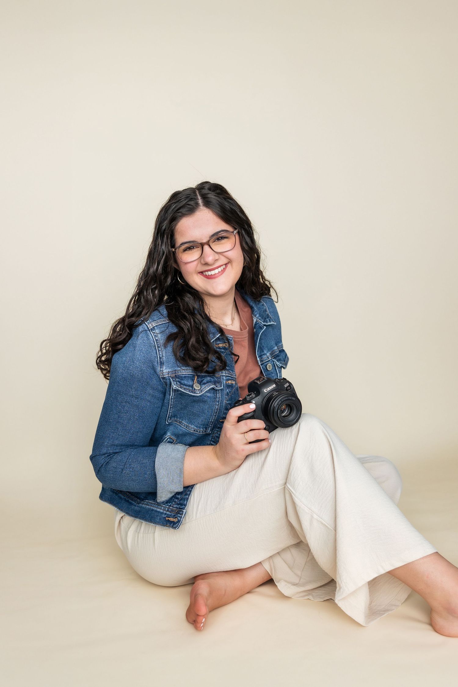 Professional photographer wearing denim jacket and glasses poses with camera against neutral background.