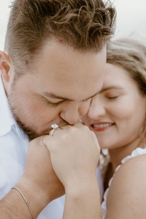 Groom kissing bride's hand showcasing diamond engagement ring during romantic beach session.