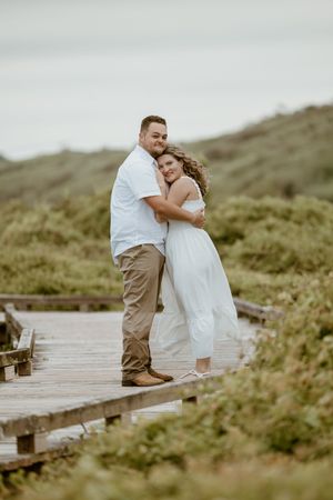 Happy engaged couple embracing on a wooden boardwalk with scenic green hills behind them.