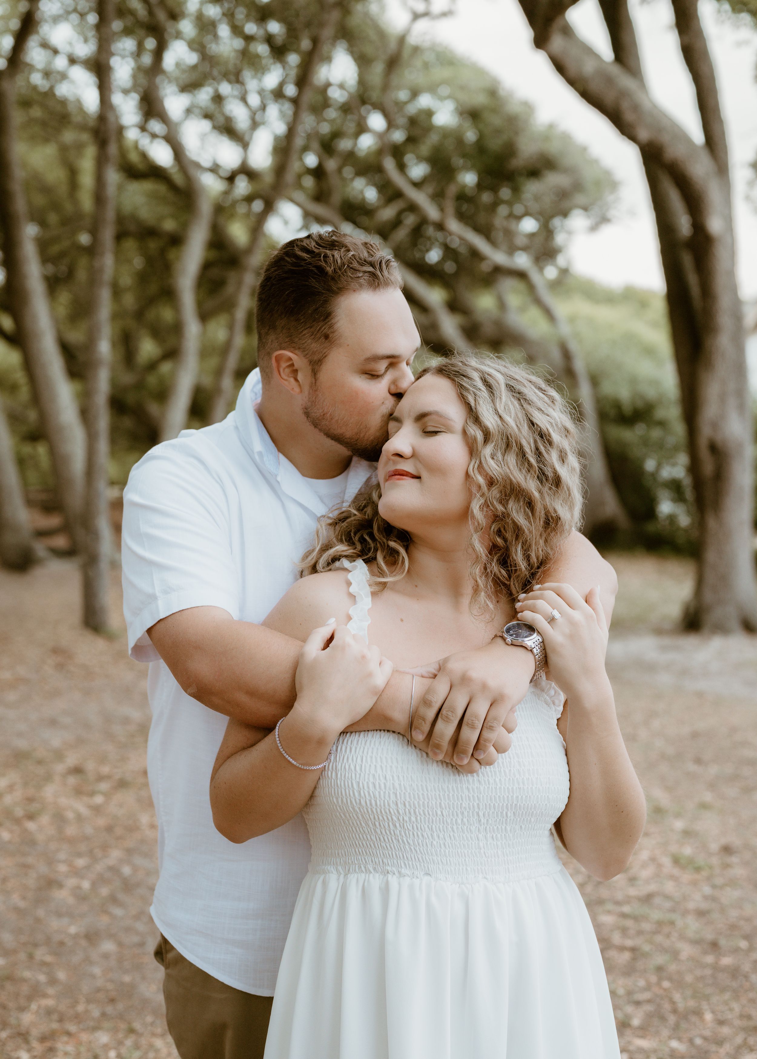 Couple sharing a tender embrace among tall trees during outdoor engagement photo session.
