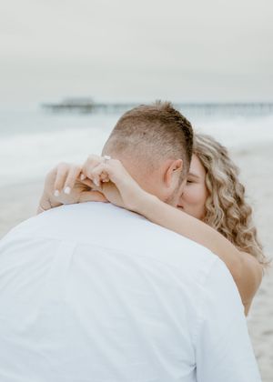 Engaged couple hugging on a beach with a pier in the background during engagement shoot.