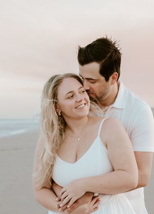 Romantic couple embraces on a sandy beach at sunset in light colored clothing, creating a dreamy coastal portrait series.