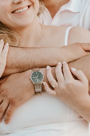 Close up of embracing couple's hands with silver watch and delicate jewelry visible in warm sunlit setting.