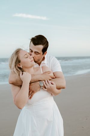 Romantic couple embracing on a misty beach while wearing white outfits against an overcast sky.