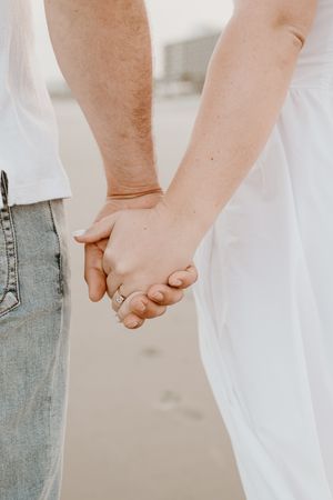 Close-up view of couple holding hands, with one person in jeans and another in white, showing engagement ring.