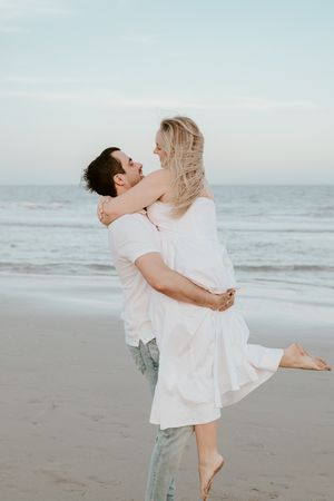 A romantic couple embraces on the beach at sunset, with the woman in a white dress being lifted by her partner.