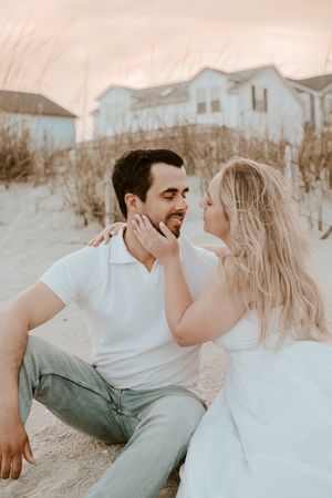 Romantic couple in white clothing share tender moments during a beach photoshoot at sunset.