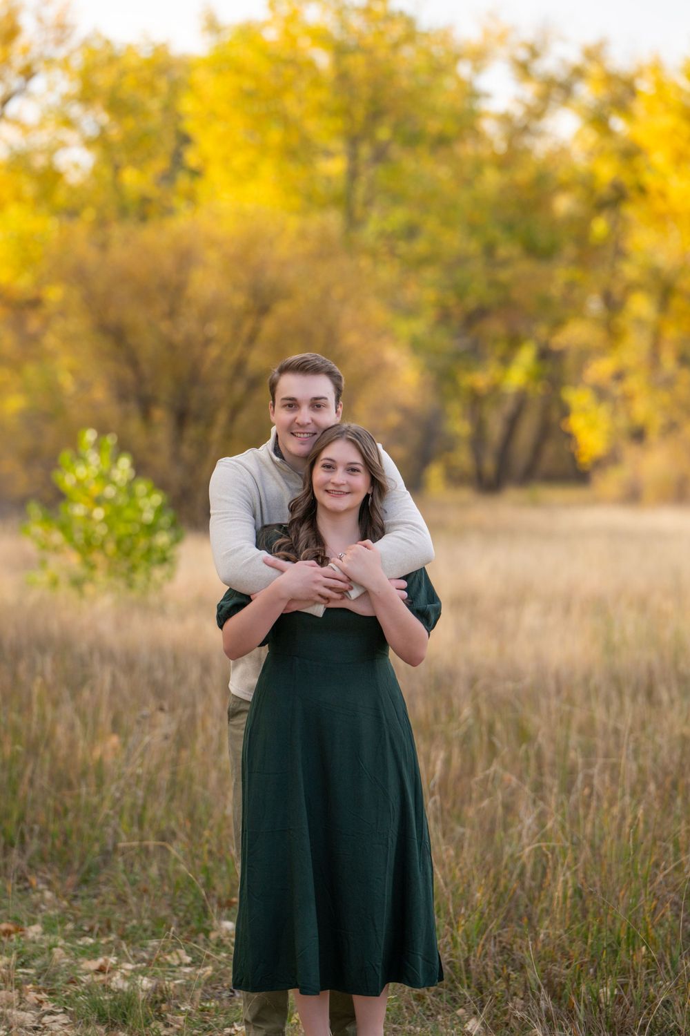 Couple embracing in autumn field with golden trees and tall grass in background.