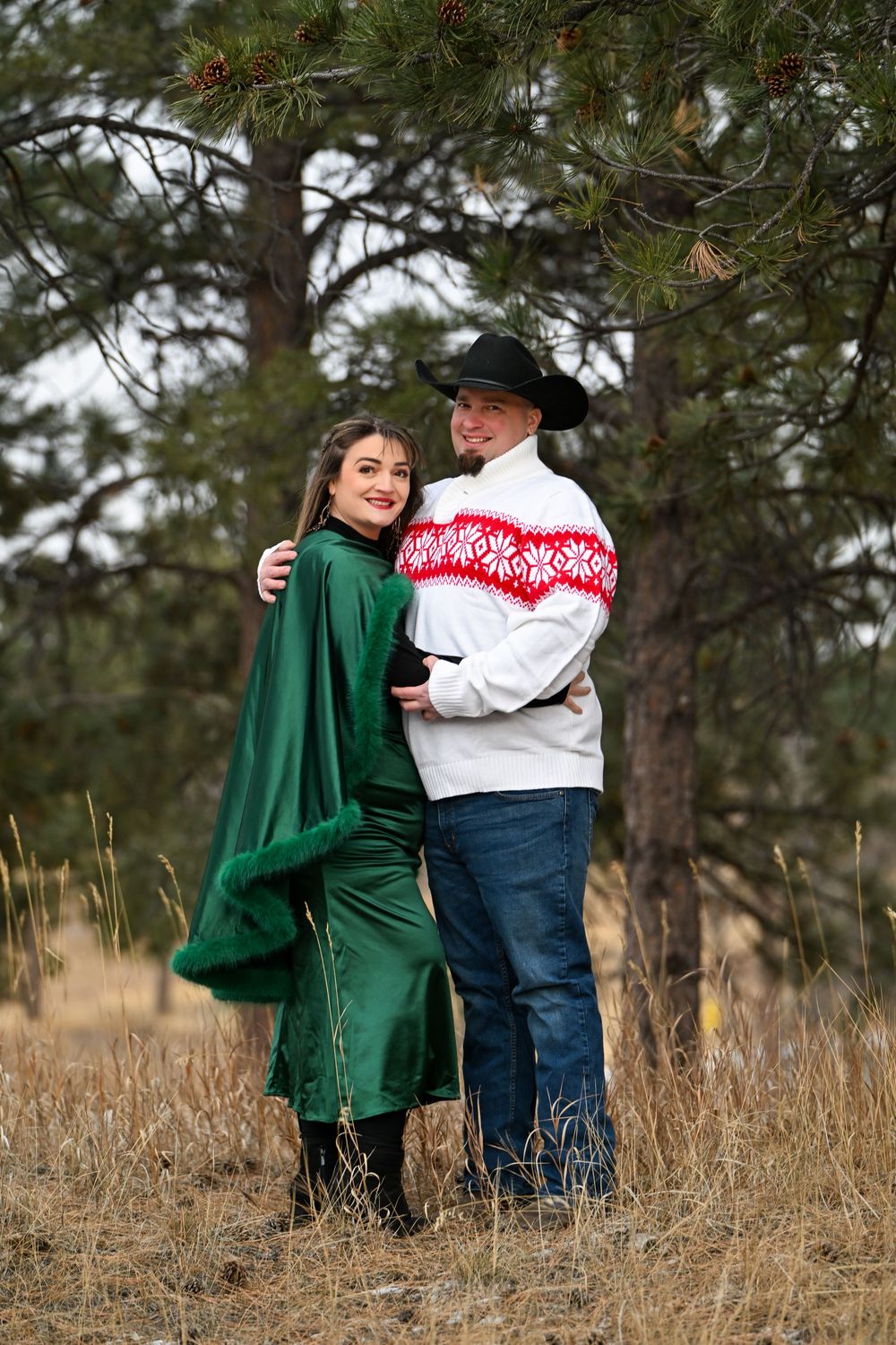 A couple wearing green and red sweaters embrace while standing in a rustic field with pine trees in the background.