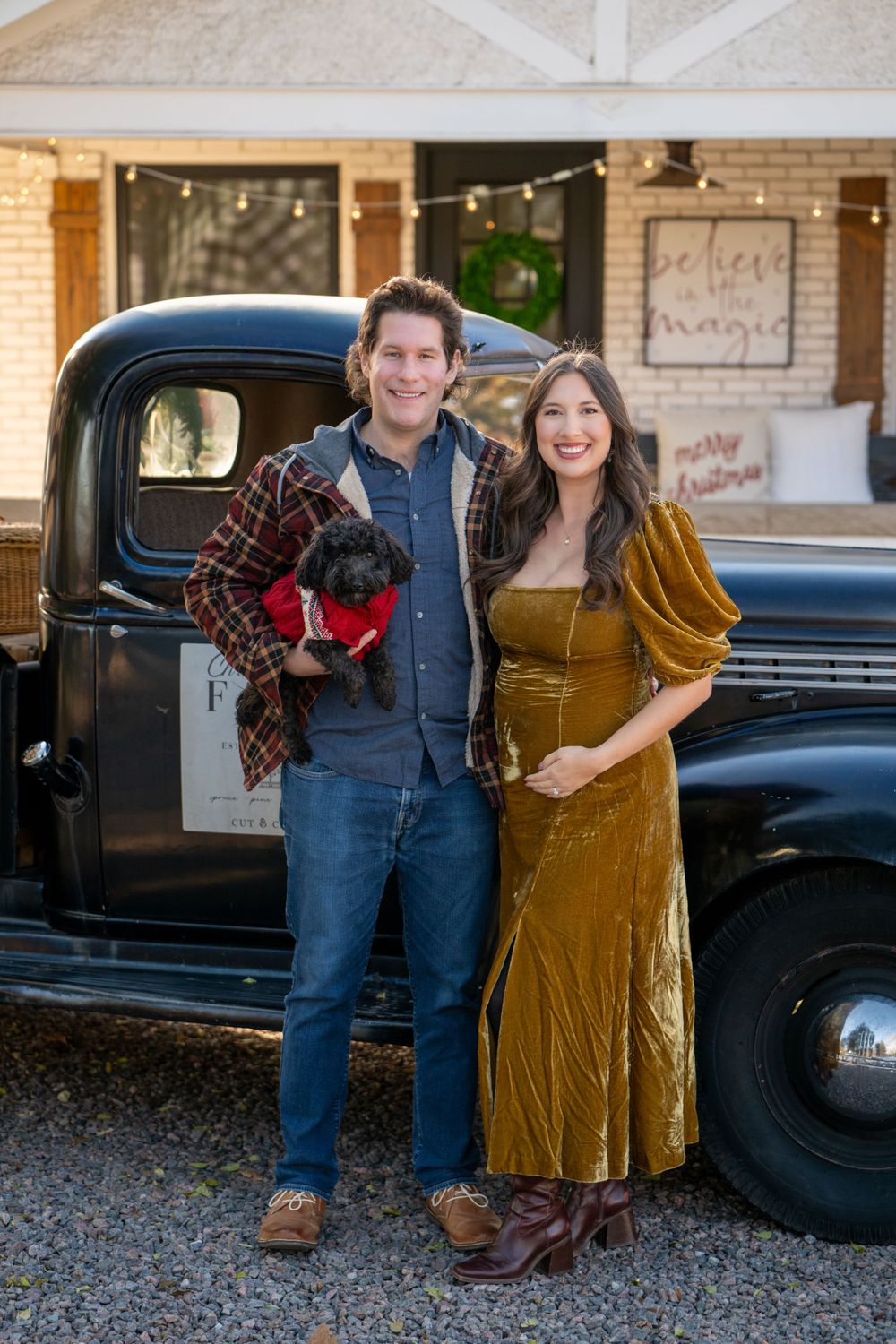 Couple poses with their black dog in front of vintage pickup truck with string lights and decorations.