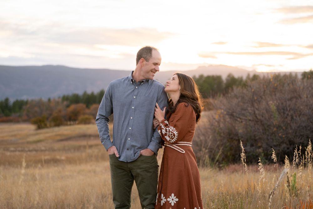 Couple in coordinating fall outfits embracing in a golden field during sunset with mountains in the background.