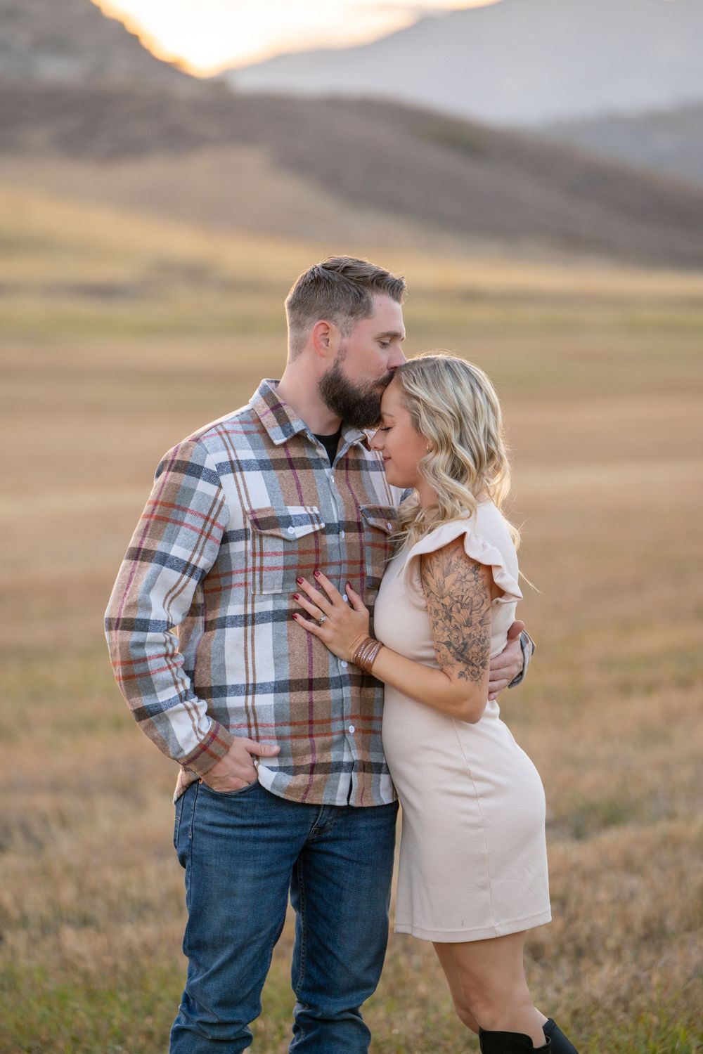 Romantic couple embracing in cream and plaid outfits against golden mountain backdrop at sunset.