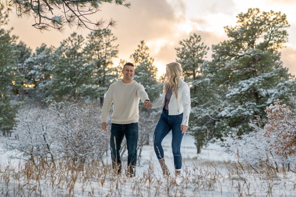 Couple walking together through a snowy winter landscape with pine trees at sunset.