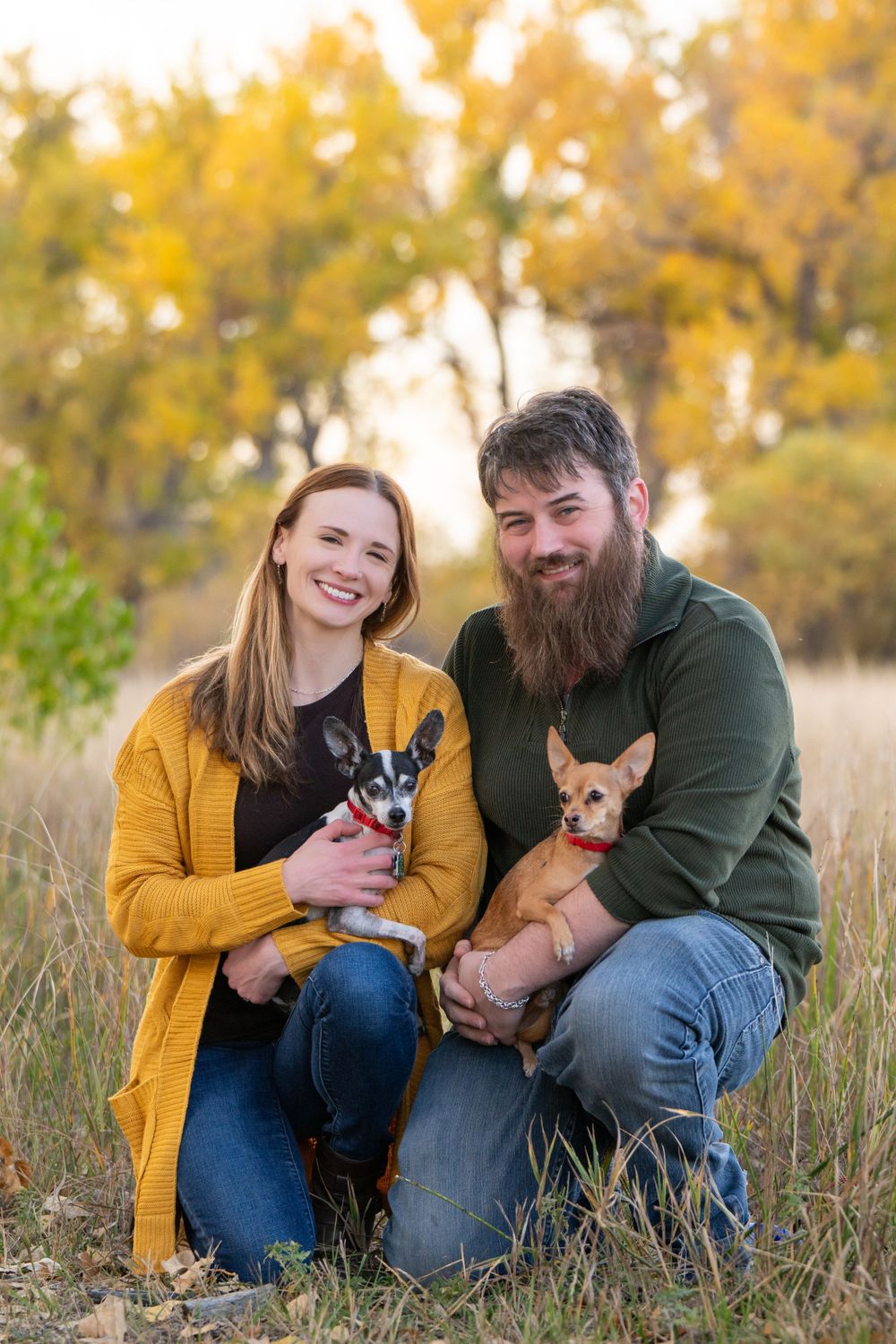 Couple kneeling in fall field holding two small dogs while wearing yellow and green casual attire.