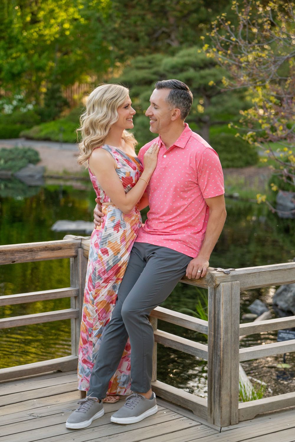 A couple stands together on a wooden bridge over a pond, wearing coordinated pink and floral outfits in a garden setting.