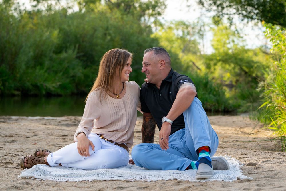 Couple sitting on white blanket by riverside on sandy beach in casual attire.