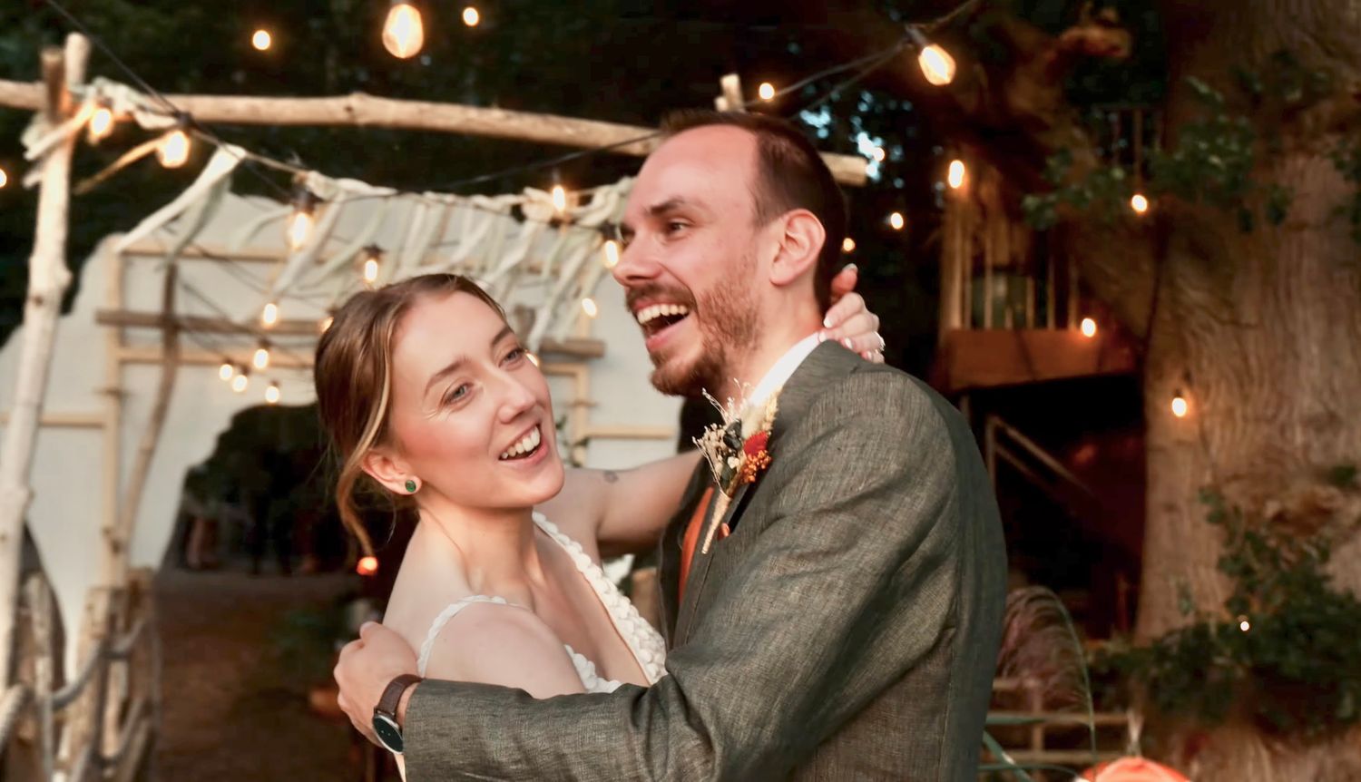 A Hollie 7 Jon, A couple laughs together under twinkling lights at a rustic outdoor wedding reception.