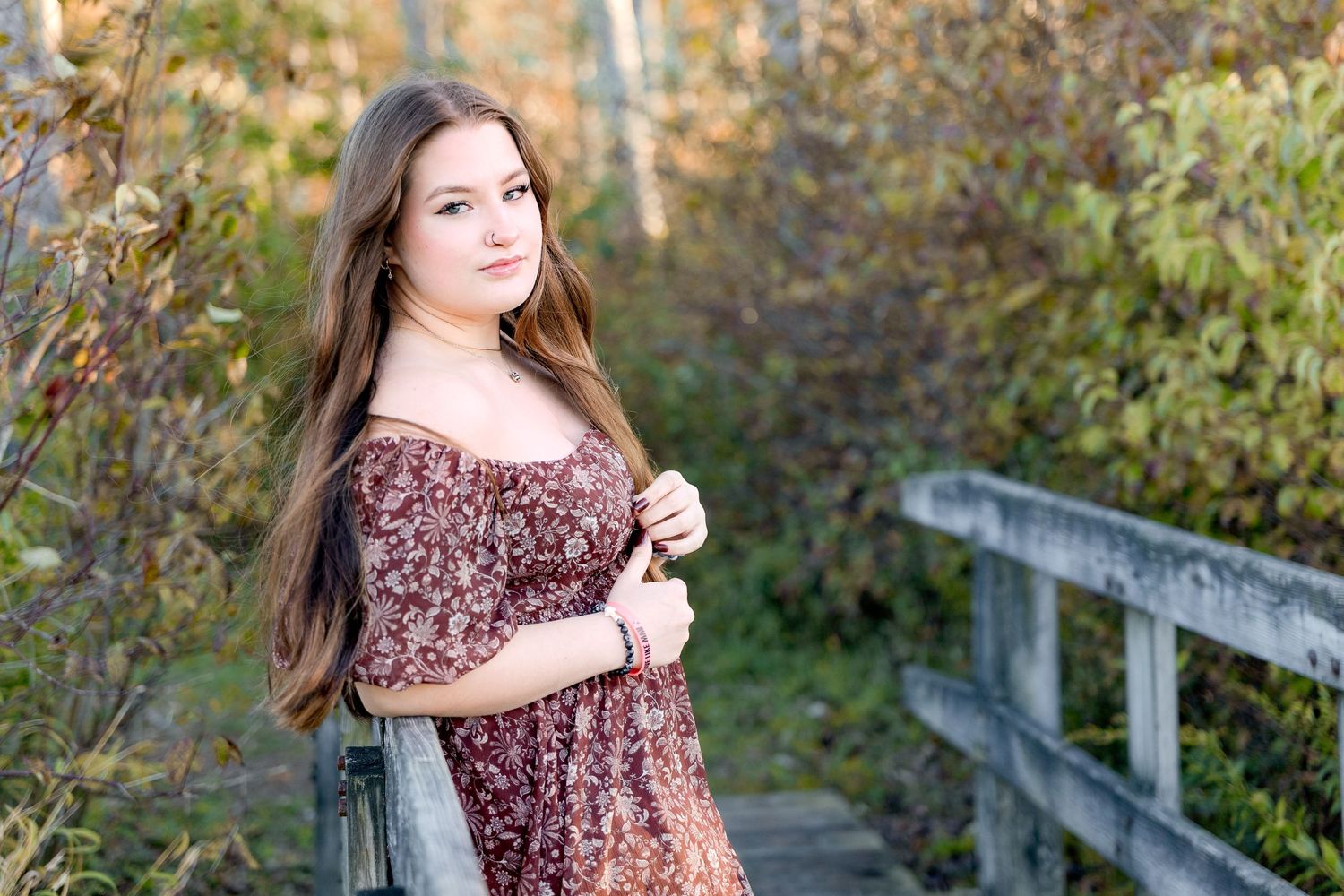 Senior in burgundy floral dress poses on wooden boardwalk during autumn season.