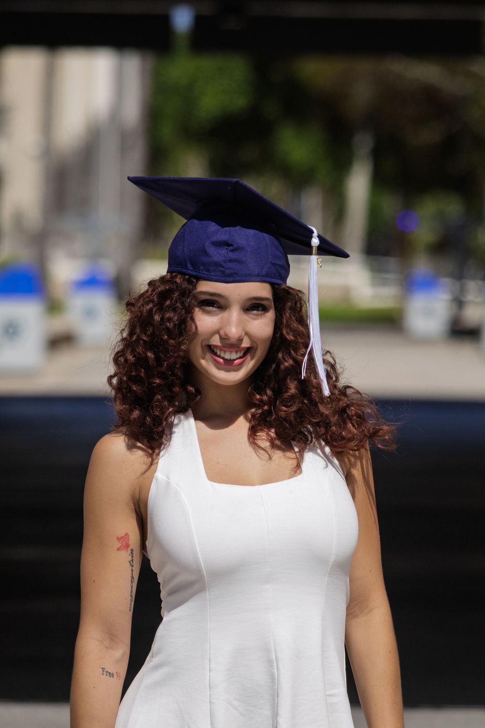 Graduate in white dress and blue cap posing outdoors on graduation day.