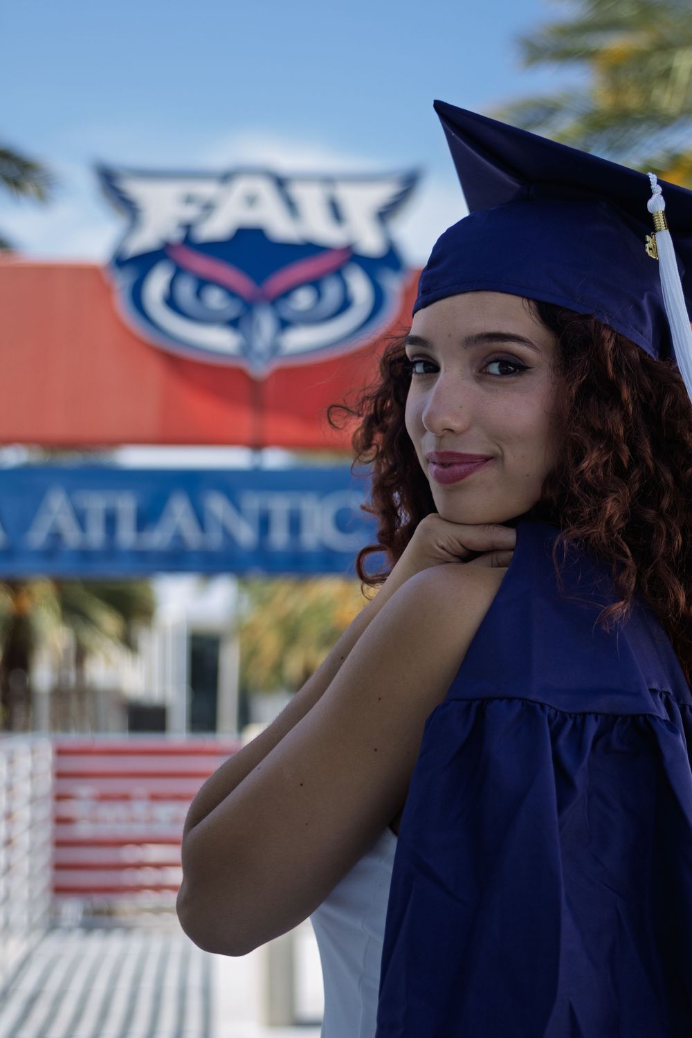 Graduate in navy blue cap and gown posing in front of FAU campus signage.