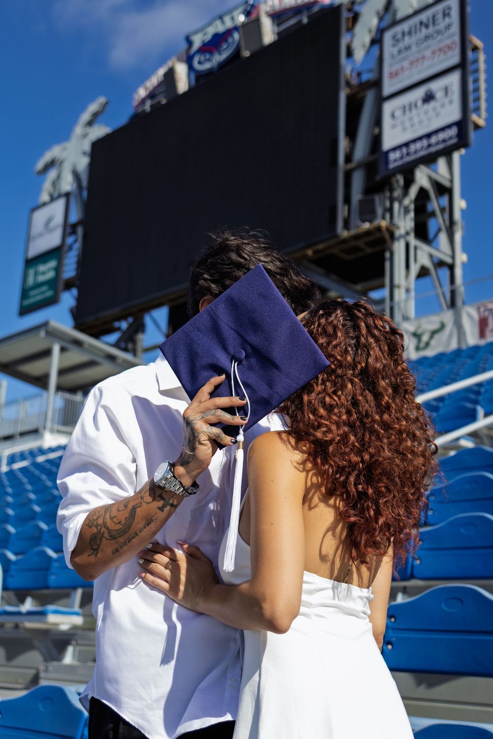 Graduate in white dress and navy cap celebrating in stadium bleachers.