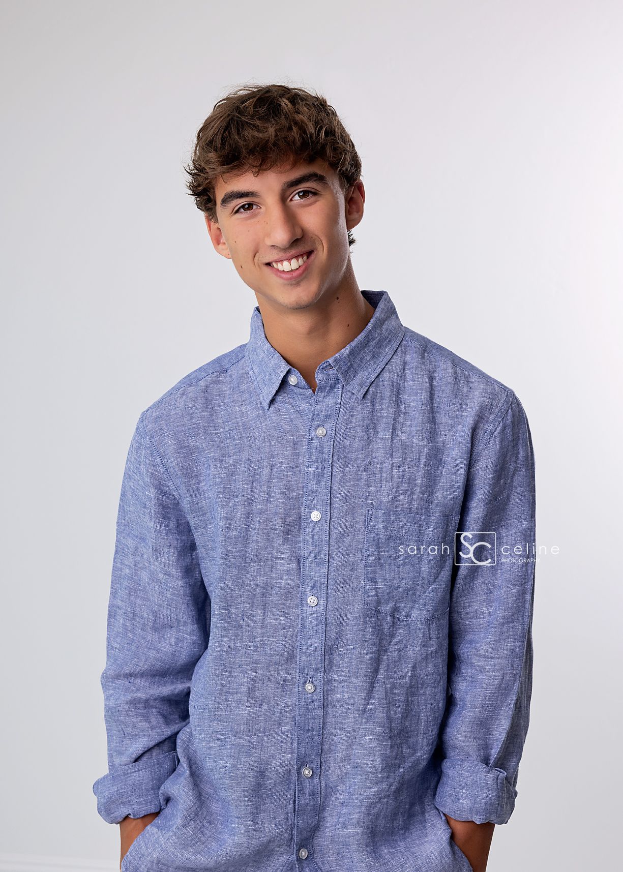 A person wearing a light blue chambray button-down shirt against a plain white background, smiling warmly at the camera.