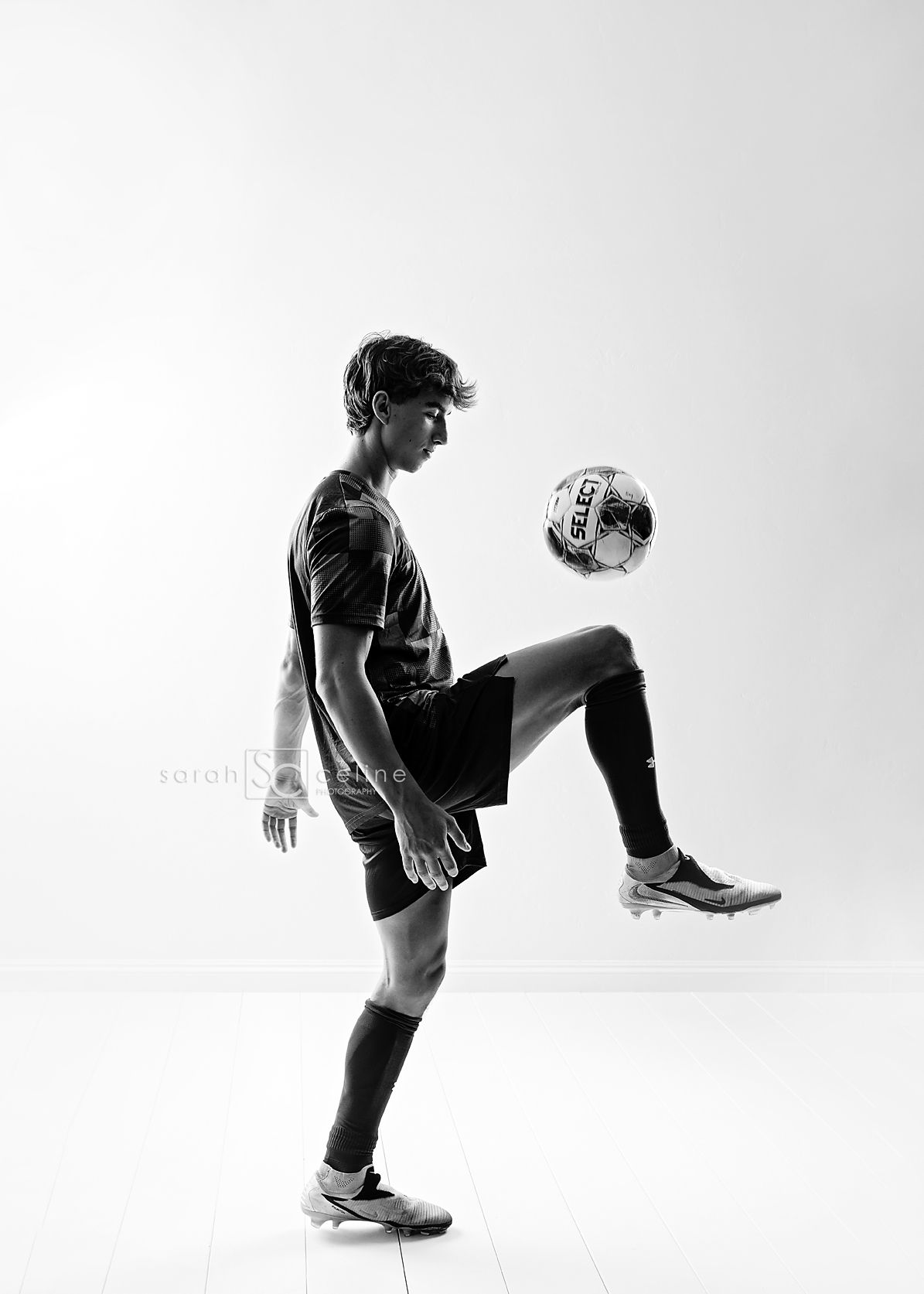 Black and white photo of soccer player juggling ball with knee in athletic pose against plain background.
