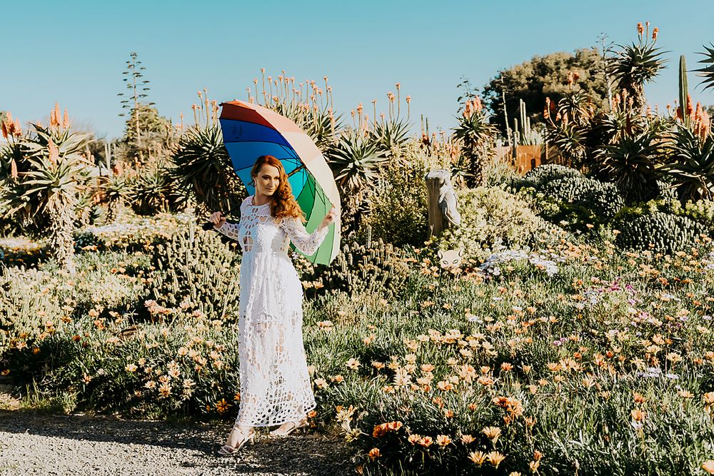 Woman in white lace dress holding colorful parasol poses in sunny garden with cacti and flowers.