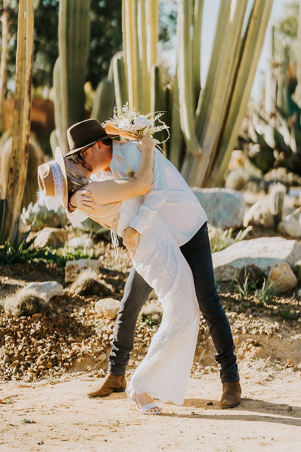 Couple in white dress and casual attire dancing playfully among bamboo trees in bright sunlight.