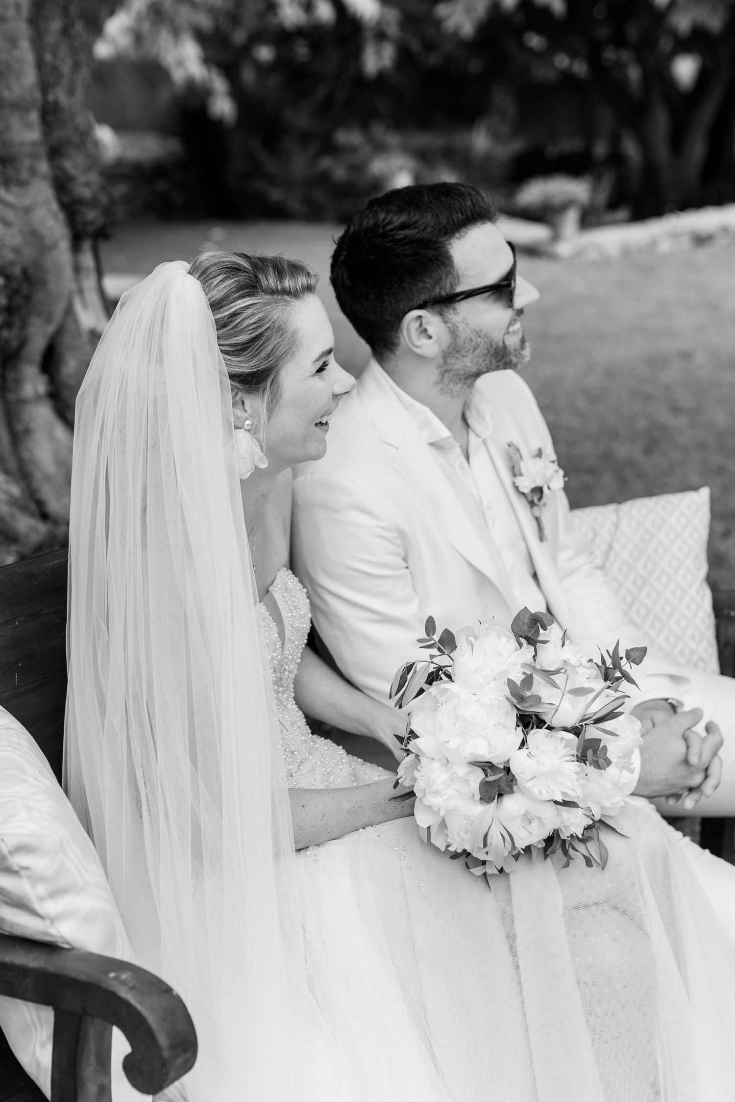 Eine Braut in einem weißen Kleid mit langem Schleier sitzt neben dem Bräutigam mit Sonnenbrille und hält einen Blumenstrauß in der Hand.